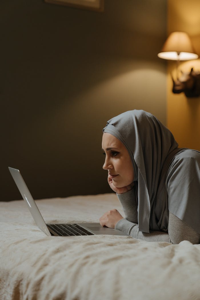 A Muslim woman in a hijab lies on a bed using a laptop, showcasing remote work in a cozy home setting.